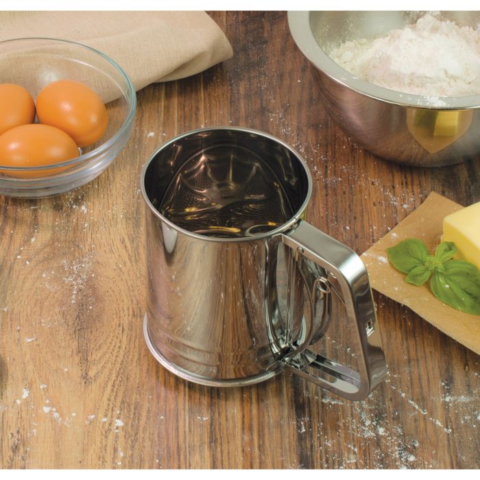 the squeeze sifter displayed with a bowl of eggs bowl of flower on a dark stained wood surface