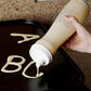 hand using batter dispenser on griddle writing "A B C" with batter.