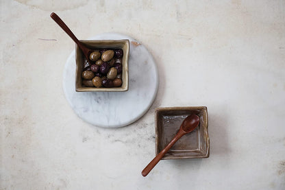 reversible roung marble cheese board displayed on a white marble surface with two square bowls one filled with olives and two wooden spoons