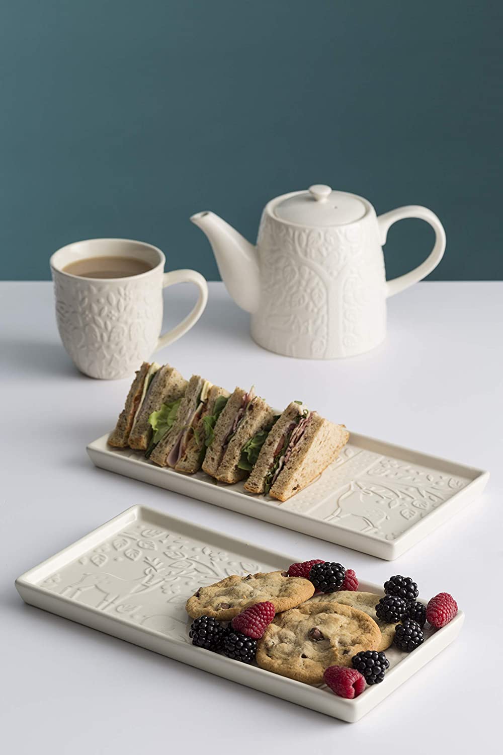 tray with cookies and berries, another tray with sandwiches, mug and teapot on white countertop.