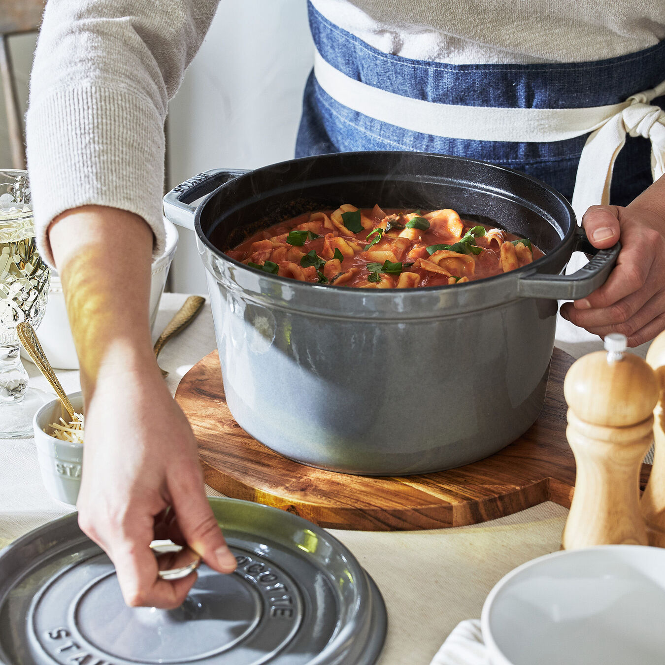 hands hold the handle and reach for the lid of a dutch oven filled with stew. scene is set on a table top with pepper mill and a glass of wine.