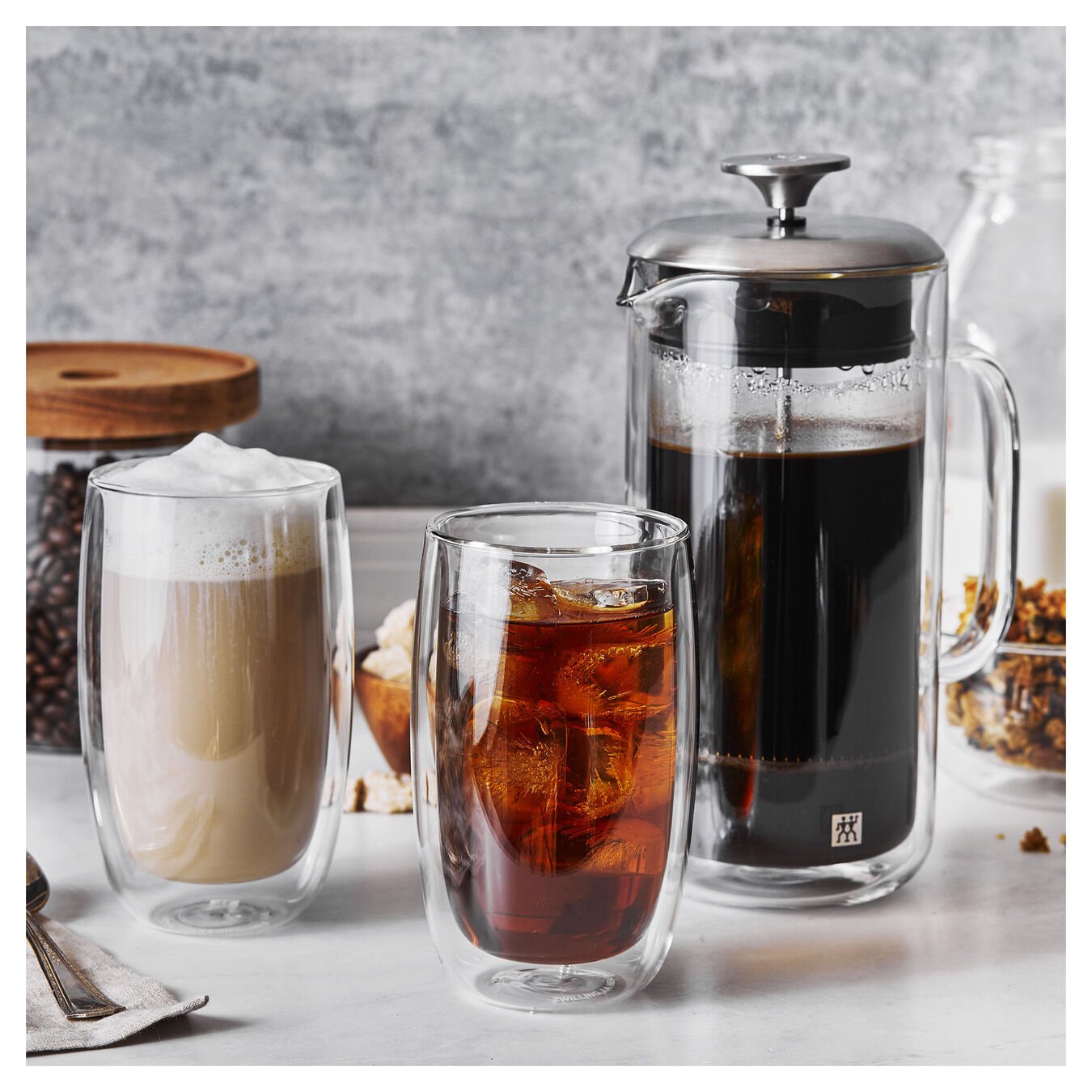 french press full of coffee and two latte glasses, one full of coffee with cream, one with an iced drink sit on white counter top with canister of coffee beans in background