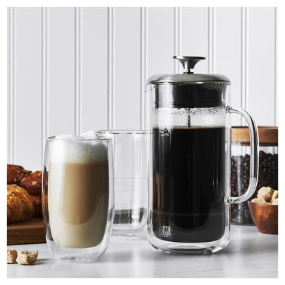 french press full of coffee and latte glasses on a white counter with white background. A canister of coffee beans and stack of croissants in the backgroundt