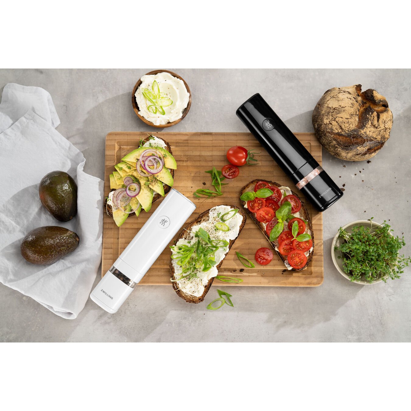 both electric salt and pepper mill displayed on a cutting board next to avocados, toast with cheese and veggies, next to a bowl of seasonings, and a white towel on a light gray surface