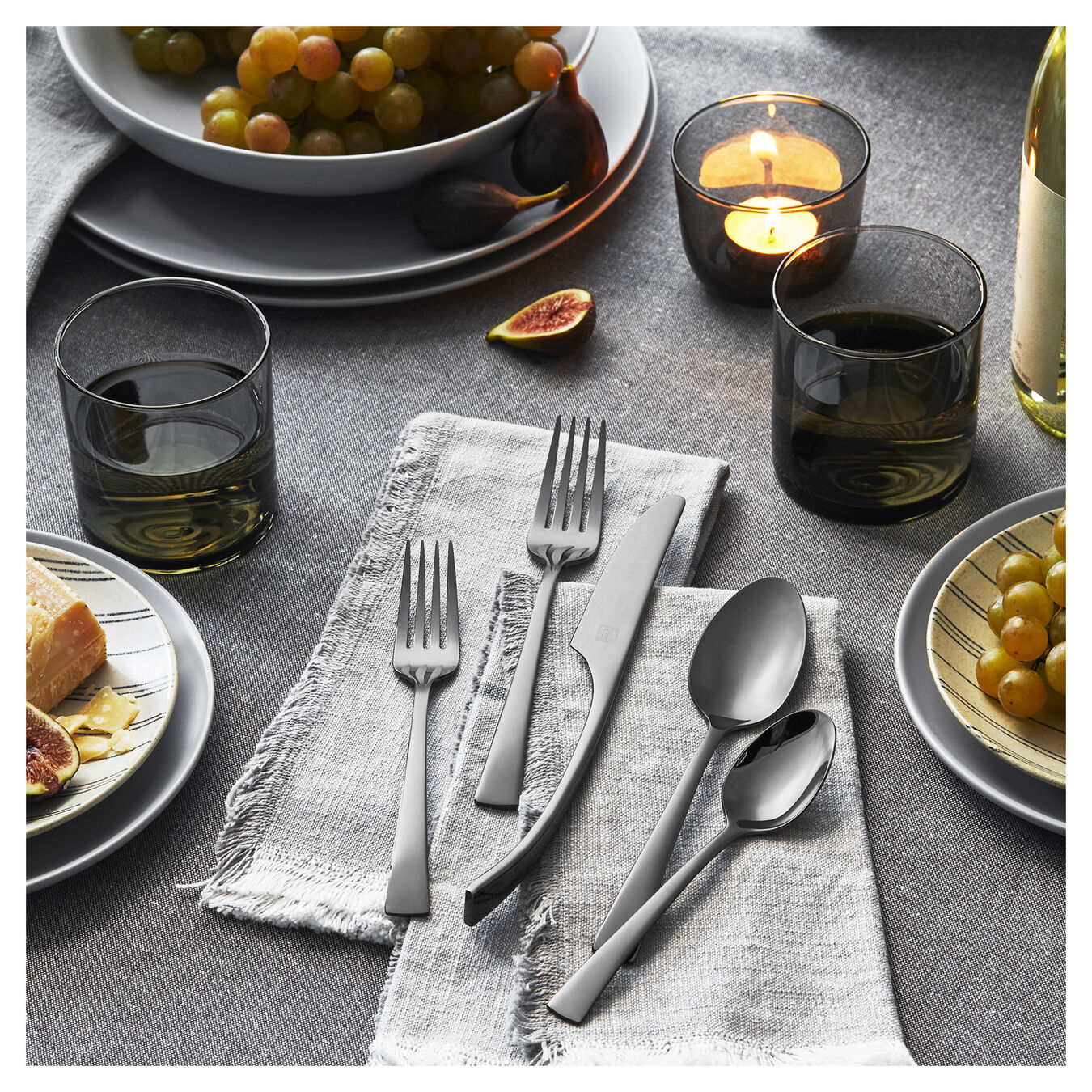 5 pieces of bellasera slate flatware displayed in a table setting next to dinner plates, drinking glasses, bowl of grapes, and light gray napkins on a dark gray placemat