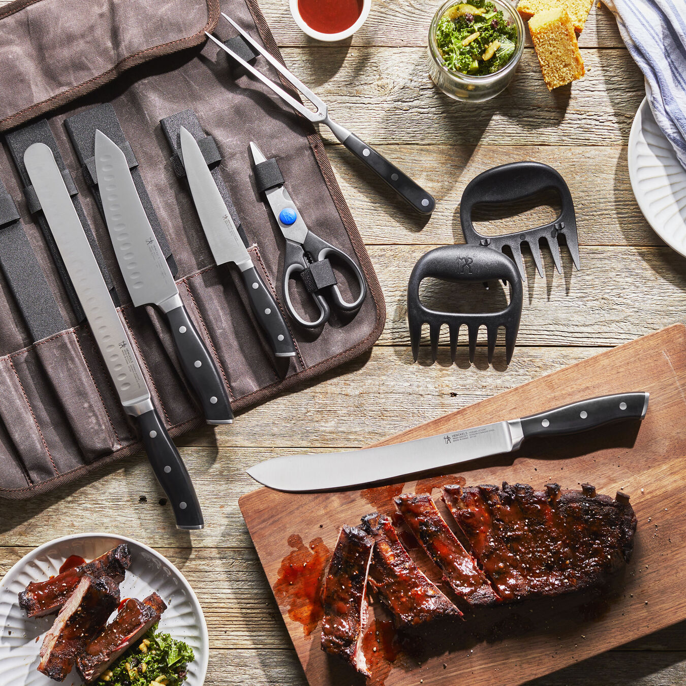 barbecue carving tool set in the roll bag displayed next to a cutting board with cooked ribs, plate of food, and bowl of sides on a rustic wood slat table