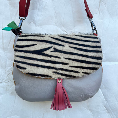 rounded grey purse with black and white striped animal print flap, pink tassel, and red strap.