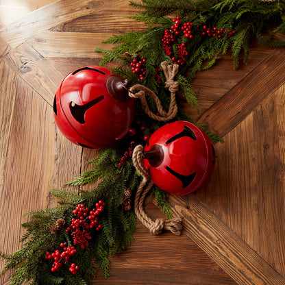 2 large red jingle bells arranged on a wooden table with greenery and red berries.