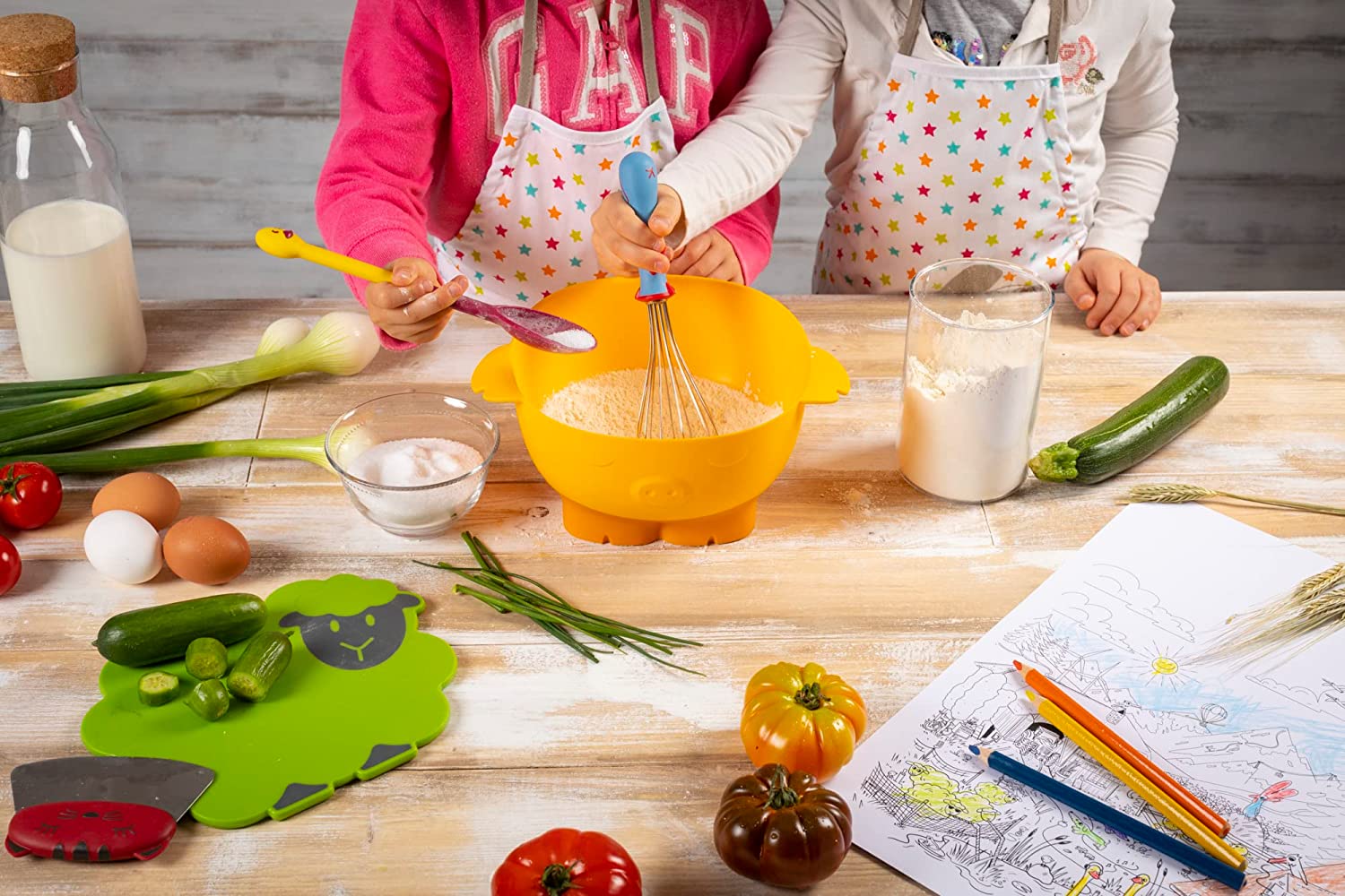 2 children whisking and spooning ingredients onto a bowl on a table with veggies, papers, and colored pencils on it.