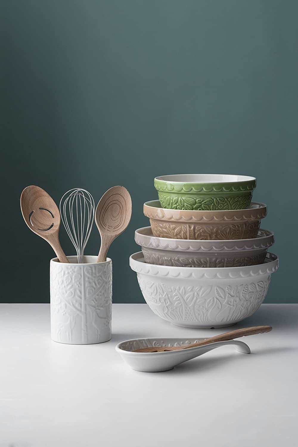 stack of bowls, crock with utensils, and spoon rest with spoon on white counter with grey background.
