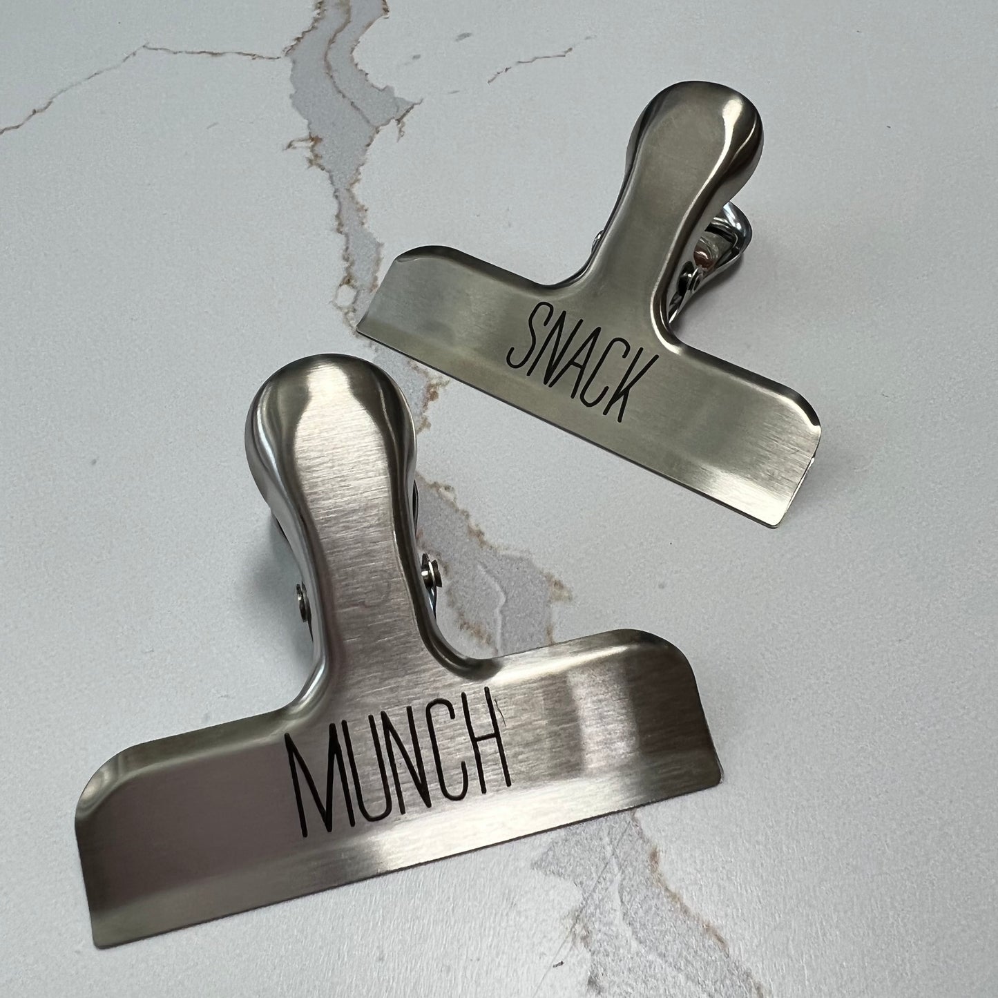"munch" and "snack" bag clips on a white marble counter.