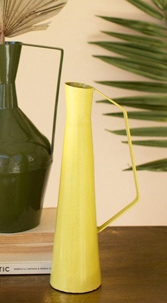 yellow metal vase with handle displayed on a wood table next to stacked books and vase against pale pink background