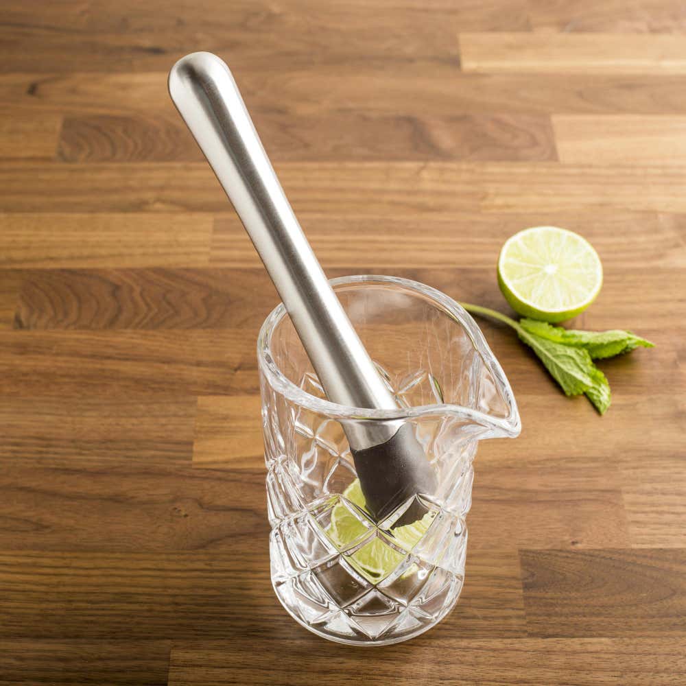 the cocktail muddler displayed in a glass sitting on a wooden surface with a lime and mint leaf