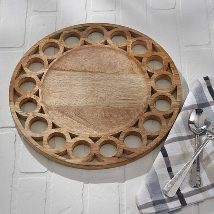round wooden charger on white brick background with plaid clith napkin and silverware on the side. charge has a ring of cut out circles around the edge and a solid circle in the center.