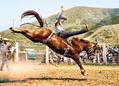 front of photo has a cowboy riding a busting bronco in a arena 