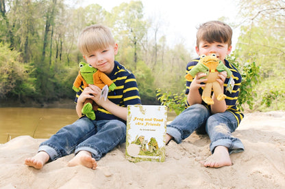 two children hold plush frog dolls sitting on a sandy beach near a river with the frog and toad book between them.