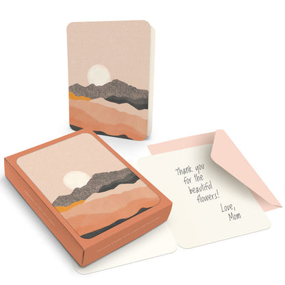 box of cards with a card and envelope outside the box on a white background. cards are blush pink with a mountain landscape and the sun behind the highest ridge. envelopes are blush pink.