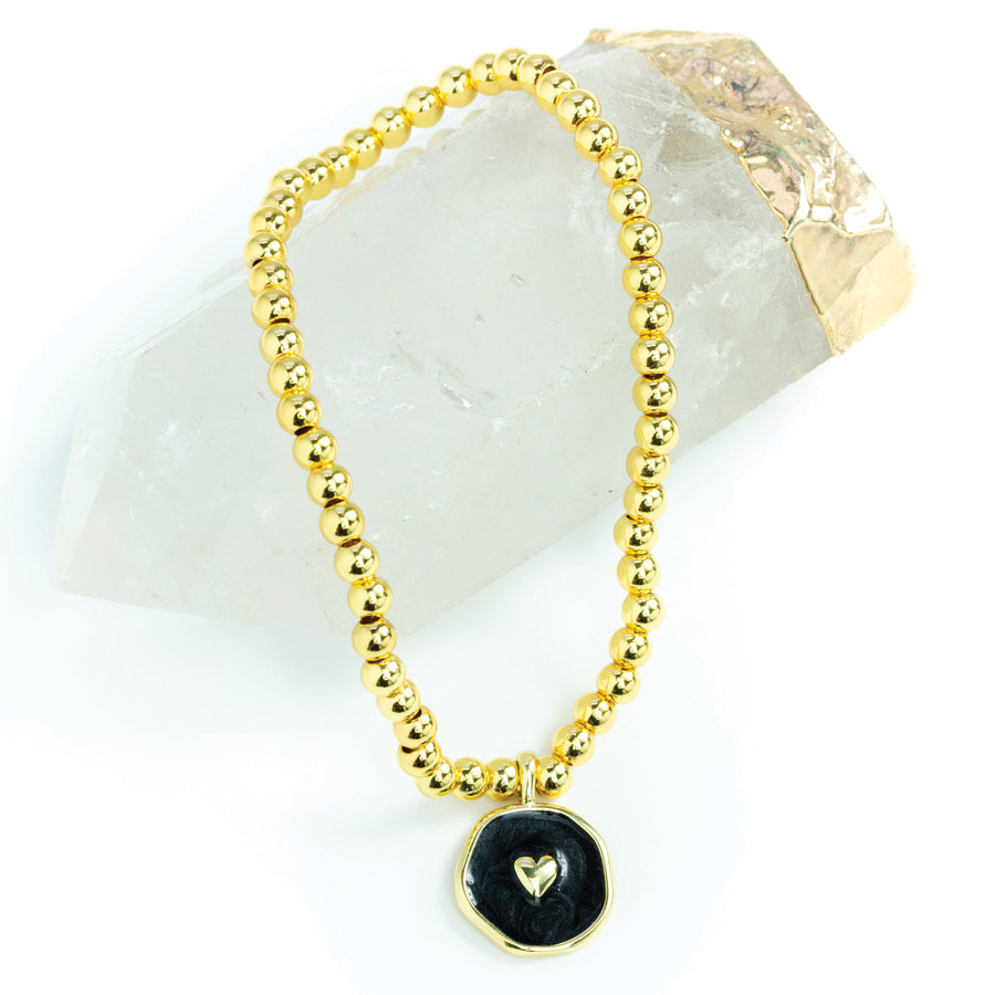 GOLD BEAD BRACELET WITH BLACK PENDANT WITH GOLD HEART ON IT LAYING ON A CRYSTAL.