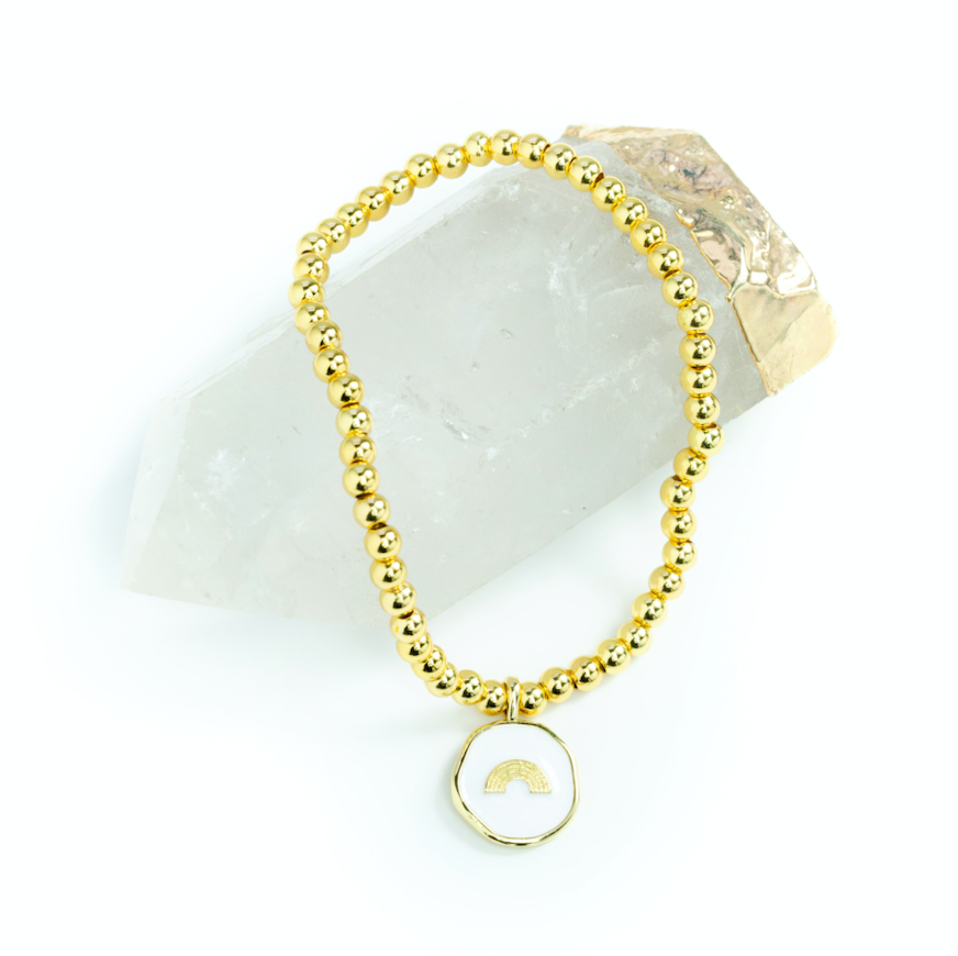 gold bead bracelet with white enamel pendant with rainbow on it laying on a crystal.