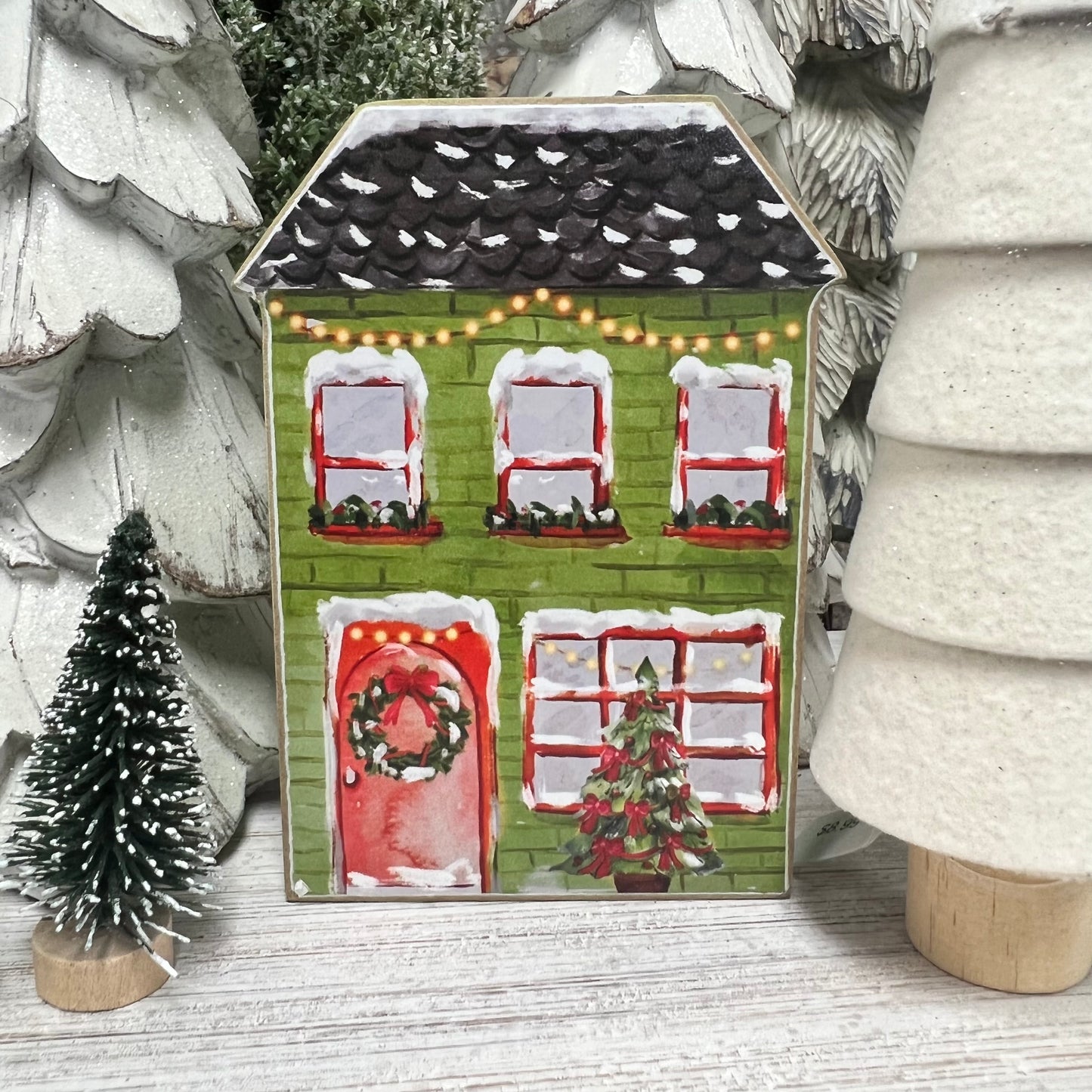 green house painted with red window trim and red door with wreath on it and lights across the top with trees set next to it.