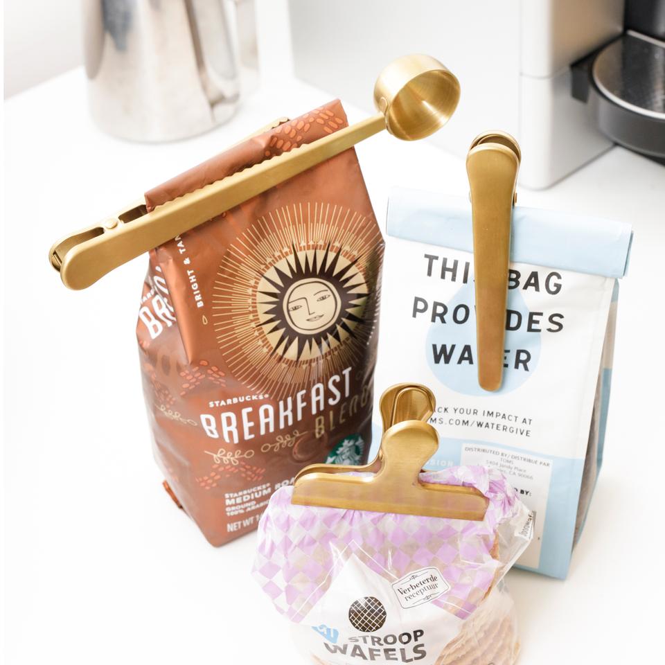 three different brass clips displayed on coffee bags next to a coffee pot on a white countertop