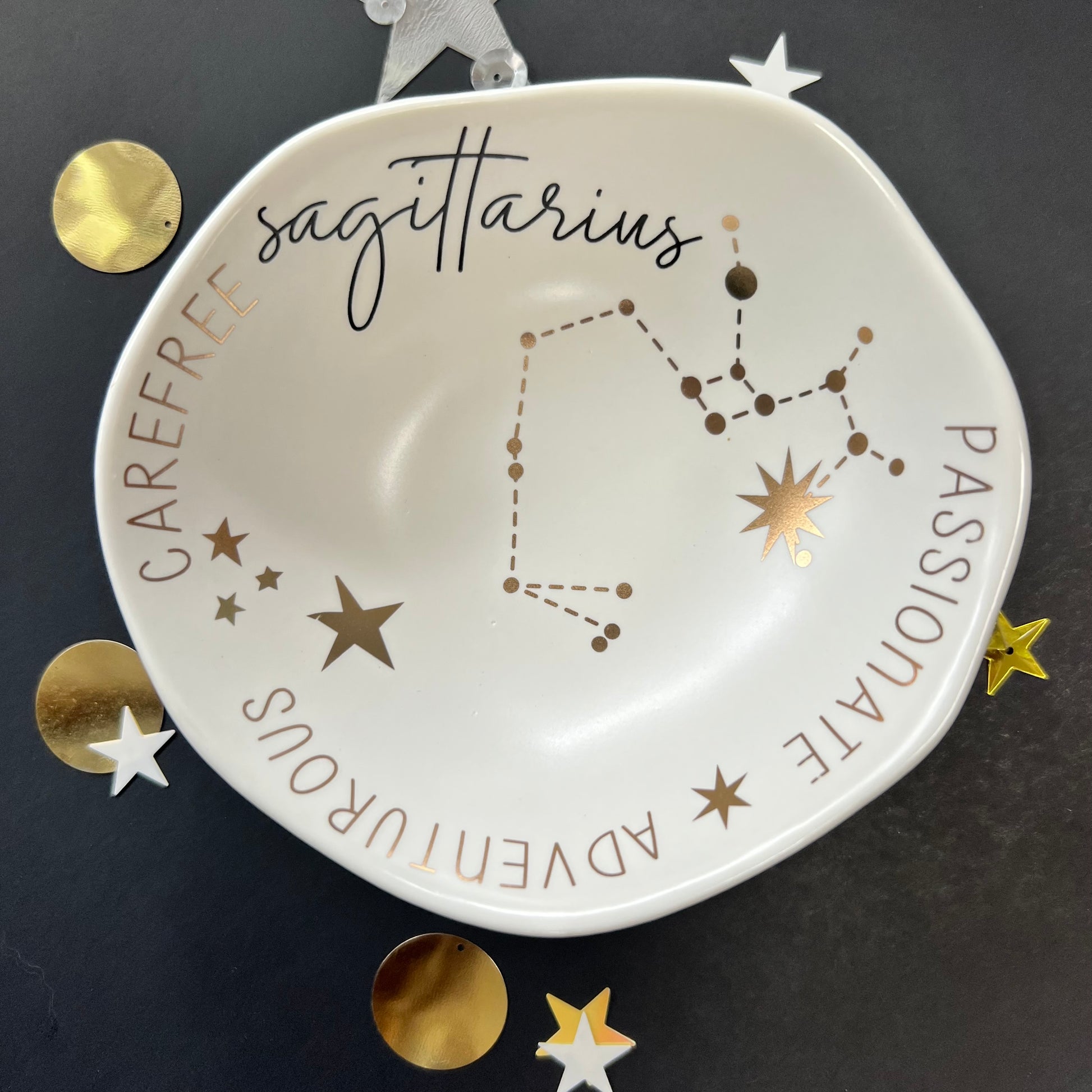cream dish with gold stars and "Sagittarius Passionate Adventurous Carefree " around the inner rim on a black background with scattered stars and orbs.