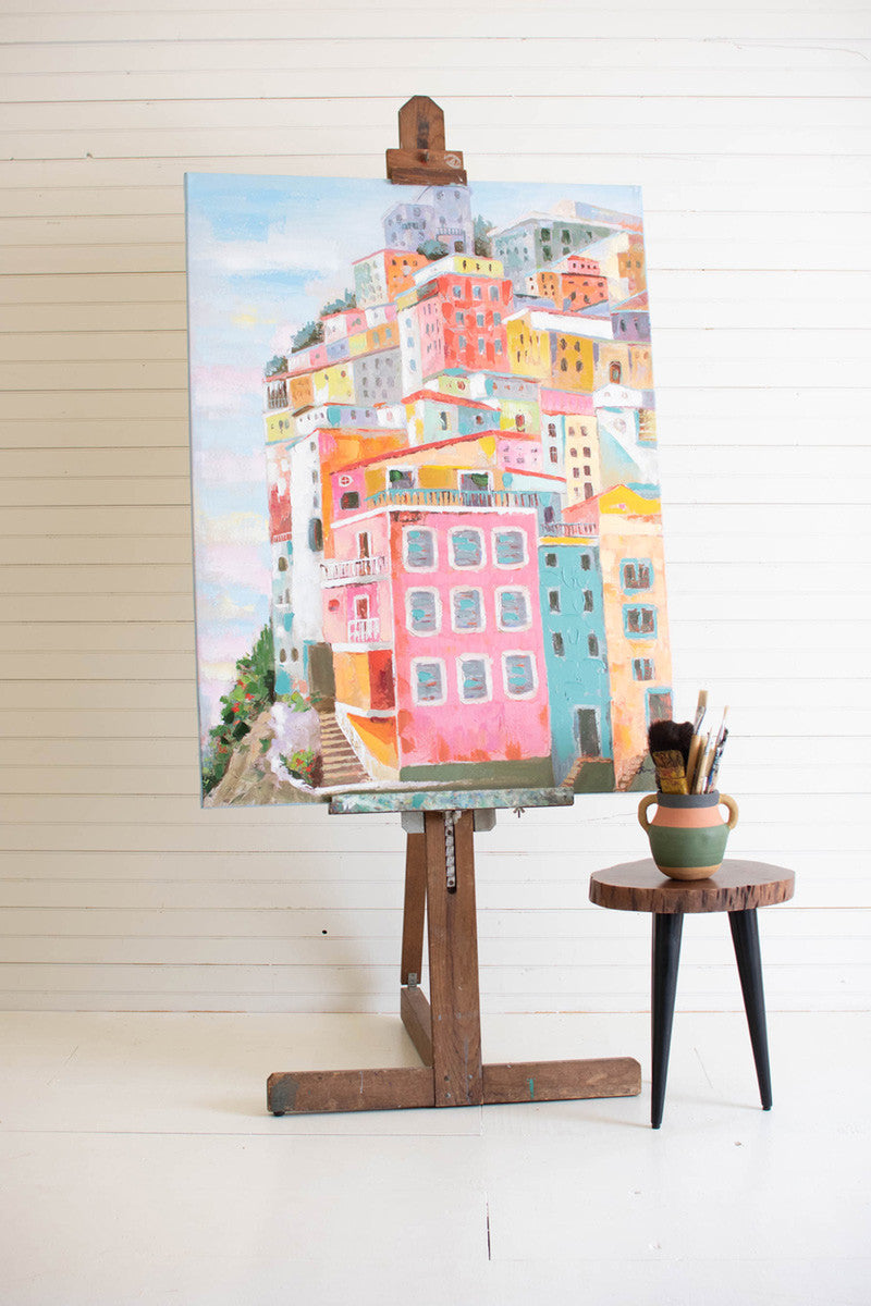 colorful houses oil painting displayed on a wood easel next to a small round table with paint brushes against a white slatted wall