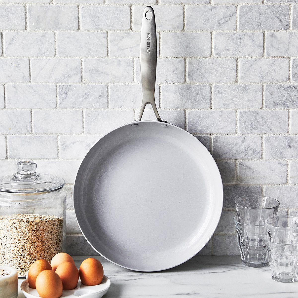 fry pan leaning on grey tile backsplash with eggs, canister, and glasses on countertop.