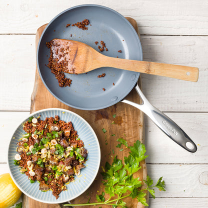 fry pan with crumbs and a spatula in it on wood board with plate of food next to it.