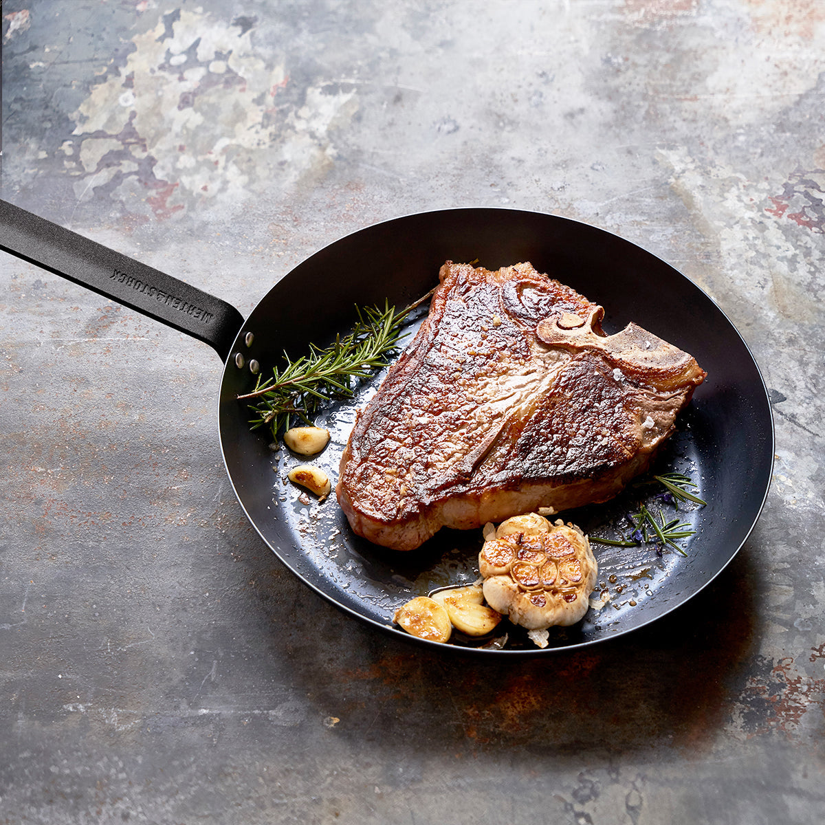 12 inch carbon steel frypan displayed with a cooked steak, roasted garlic, and seasoning on a distressed concrete surface
