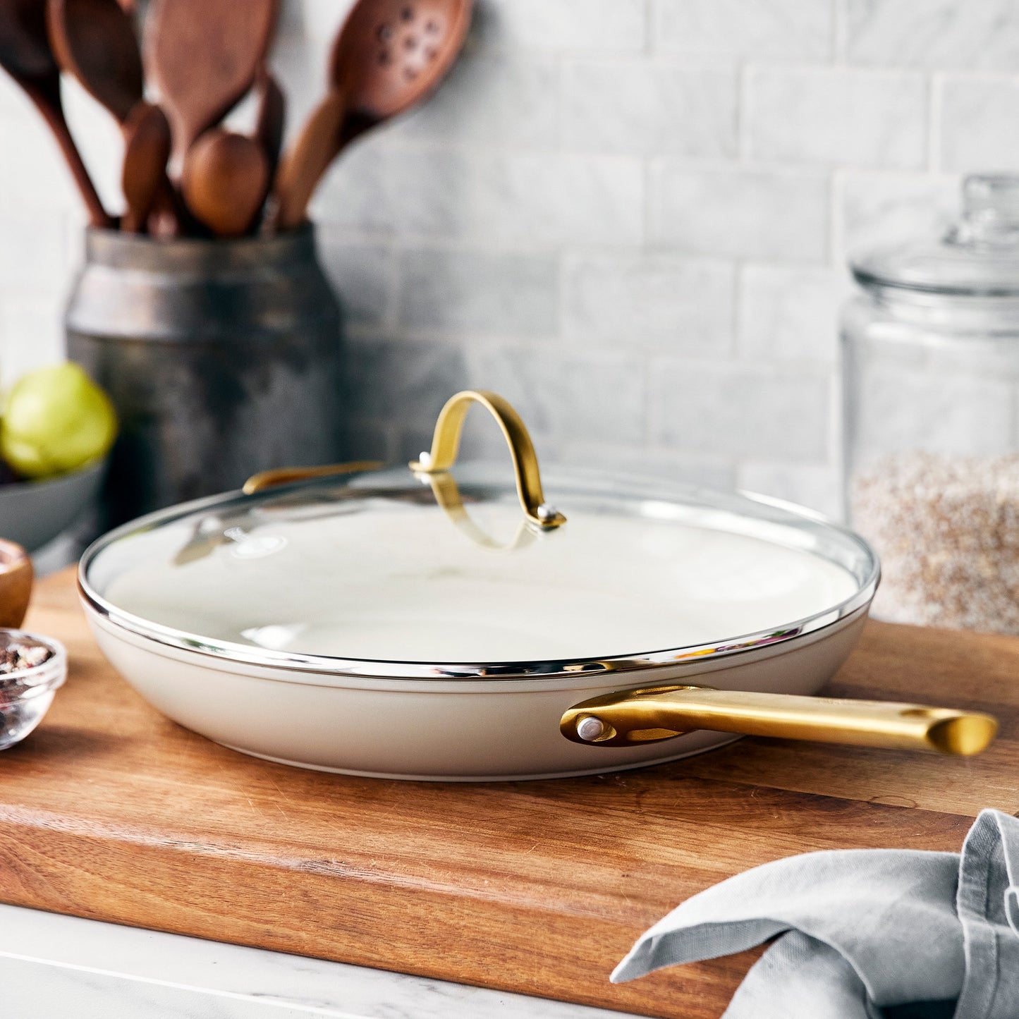 taupe reserve covered frypan displayed on a cutting board on a countertop next to a rustic utensil jar, and glass flour container