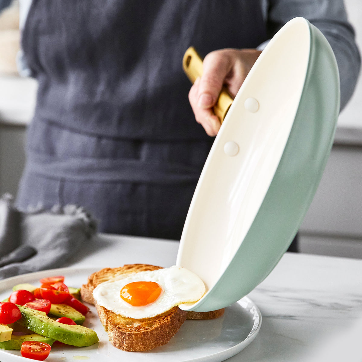 fried egg sliding out of fry pan onto plate with toast, avocado, and tomatoes on it.