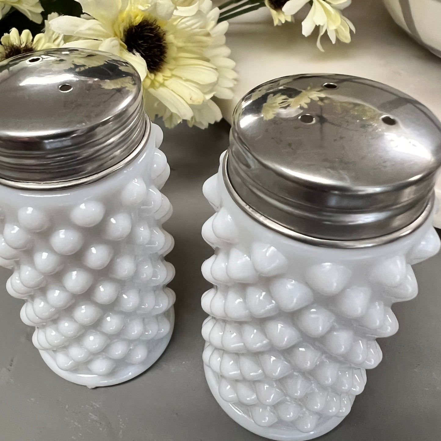 angled view of milk glass salt and pepper shakers displayed on a gray surface next to a bundle of flowers