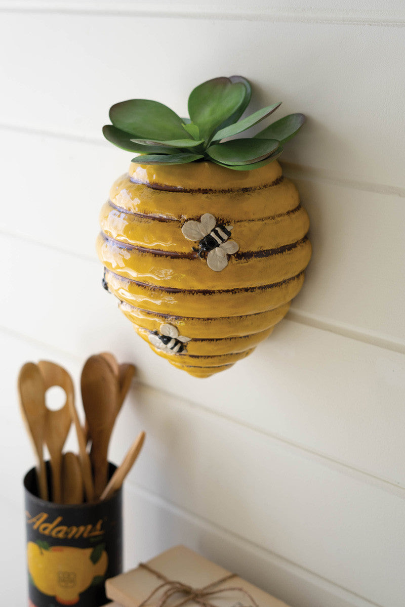 ceramic bee hive wall planter filled with a large leaf succulent hanging on a white wood slat wall above a kitchen counter with books and utensil holder filled with wooden utensils