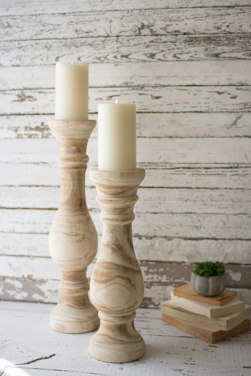 small and large carved wooden candle stands displayed with stacked books and small succulent against a rustic whitewashed slat wall