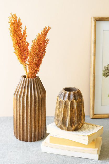 small and large carved wood vase displayed on and next to a short stack of books next to a picture and the large filled with orange stems