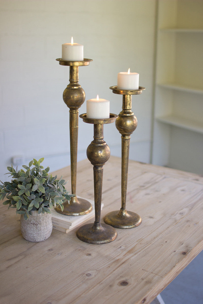 all three sizes of antique brass finish candle stands displayed next to a potted plant on a light wood table against a white wall