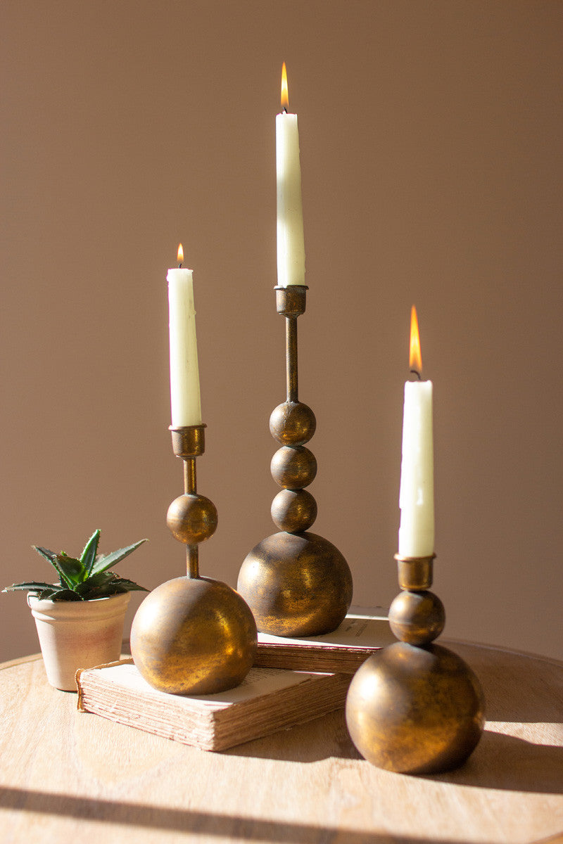 all three sizes of antique brass taper candle holder displayed with burning candles next to a succulent displayed on books against a taupe background