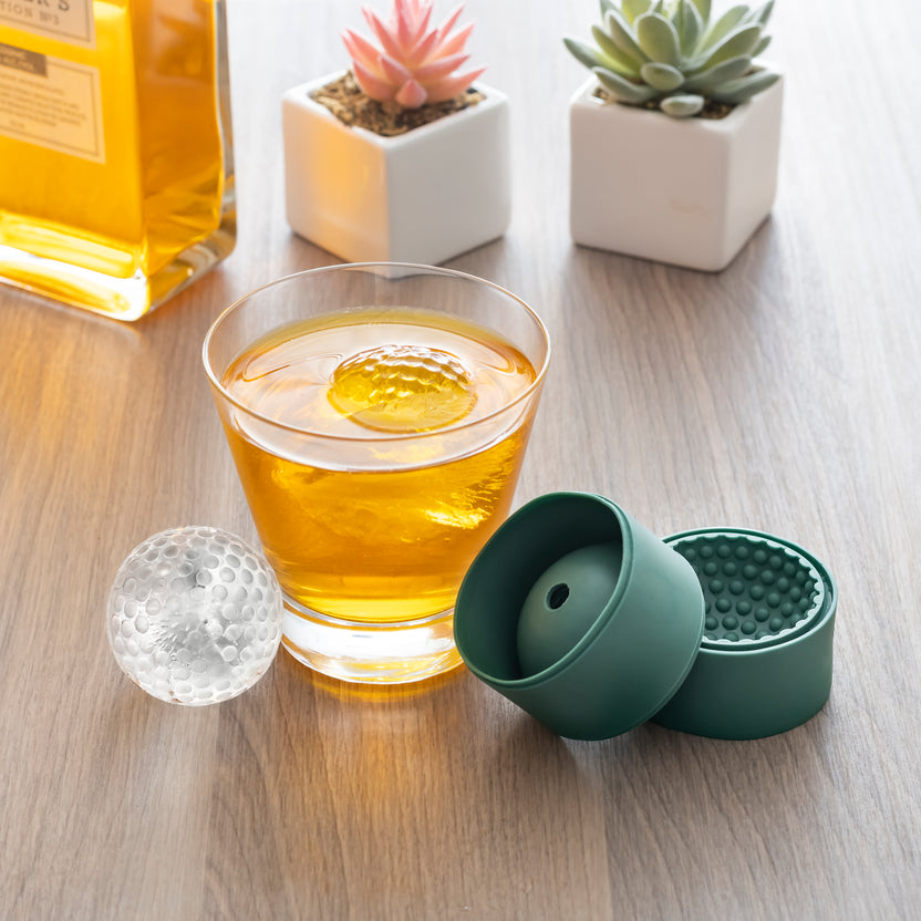 drink with golf ball ice cube in it, and golf ball ice cube and silicone mold set next to it on a table with small plants and a bottle.