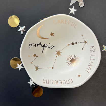 cream dish with gold stars and "Scorpio Brilliant Endearing Careful" around the inner rim on a black background with scattered stars and orbs.