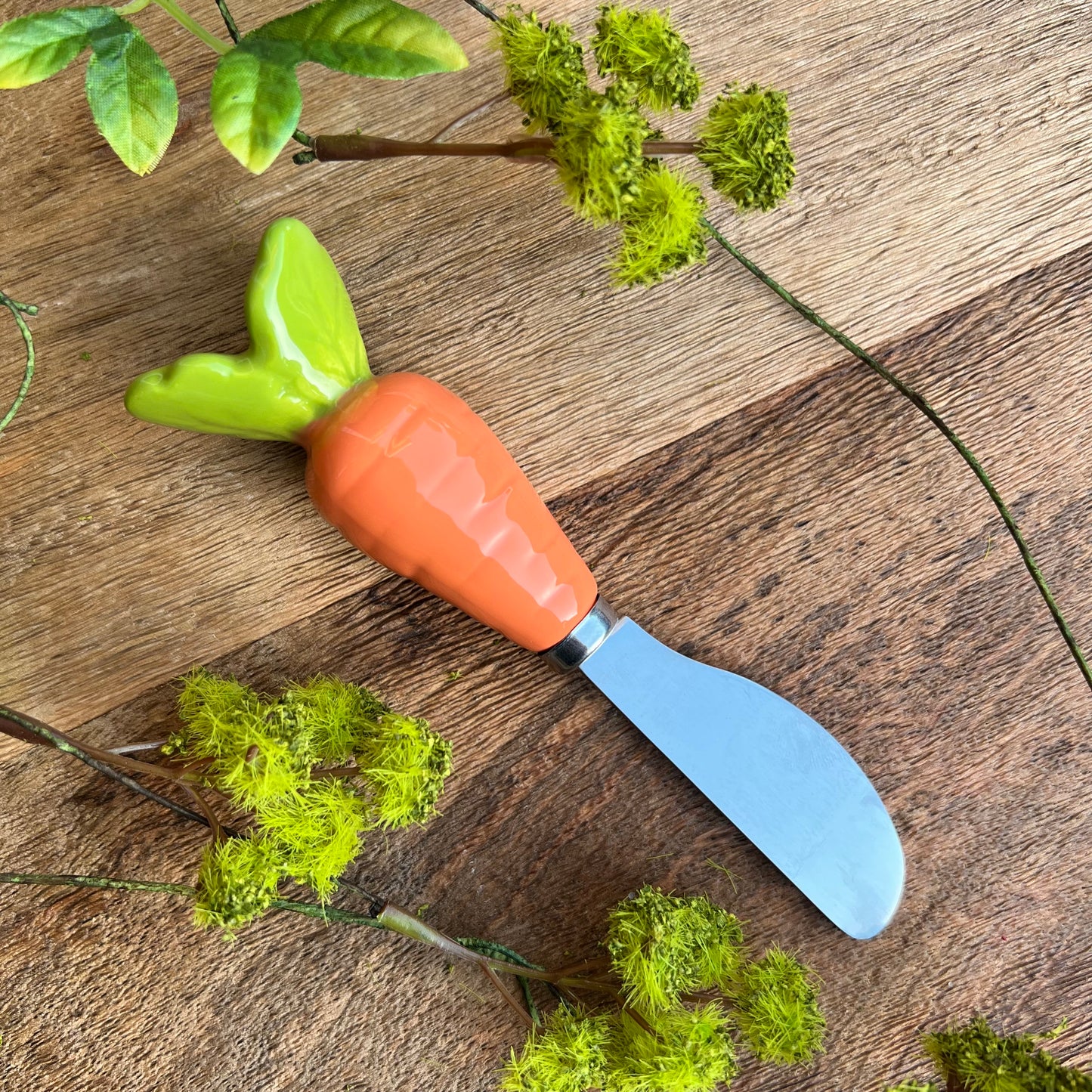 ceramic carrot handle on stainless steel spreader on wooden background with sprigs of greenery.