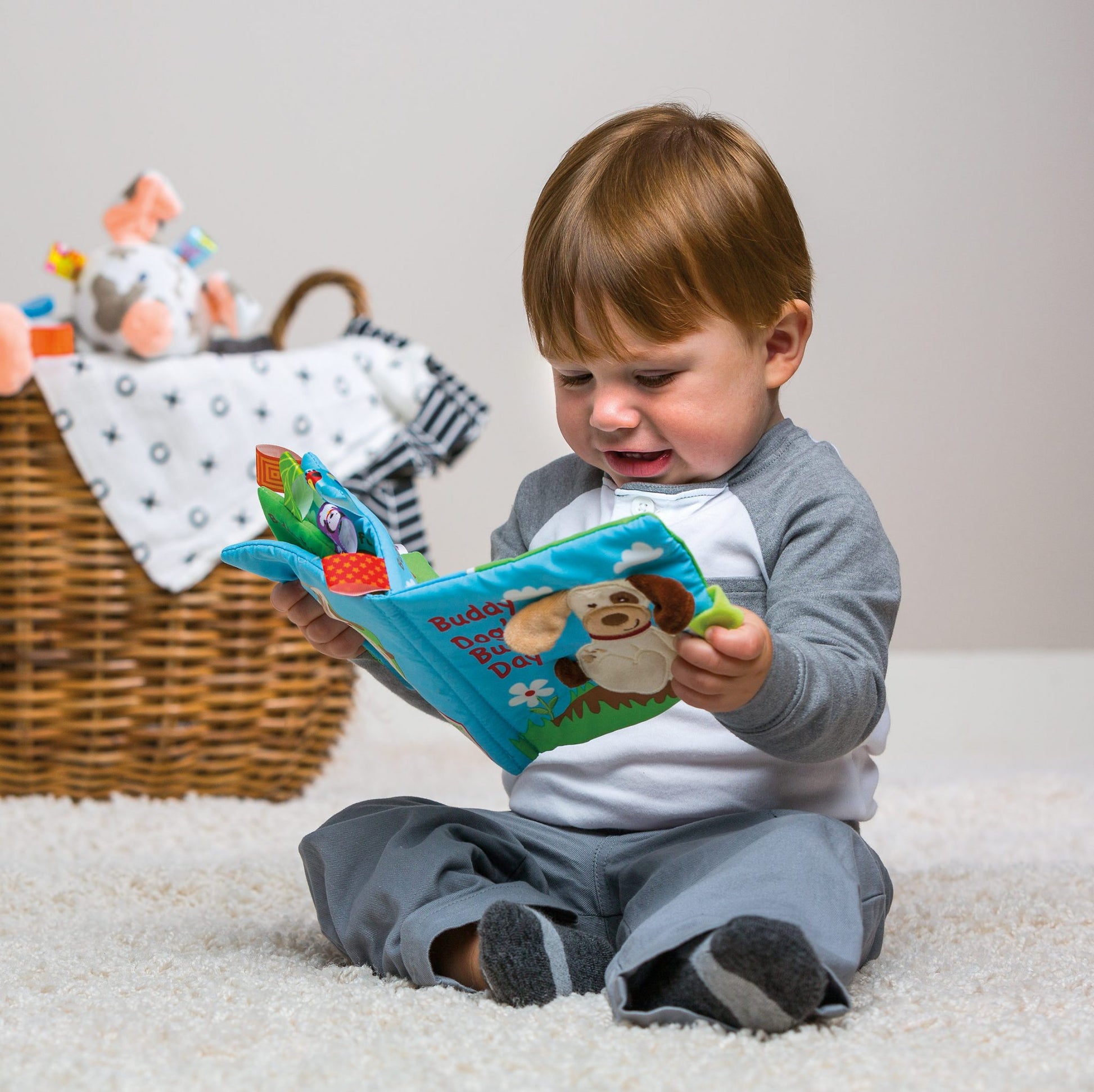 a little boy looking at the buddy dog soft book while sitting on a white rug next to a basket of toys