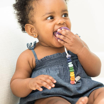 little girl sitting with a paci in her mouth and attached to the rainbow pacifier clip against a white background