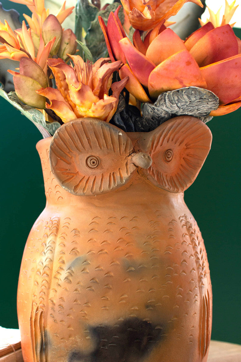 close up view of the terracotta owl planter displayed with orange floral arrangement on a light wood table with small stacked books against an emerald background