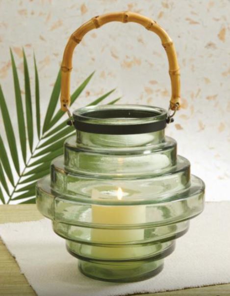 tiered glass lantern with lit candle inside set on a table with greenery in the background.