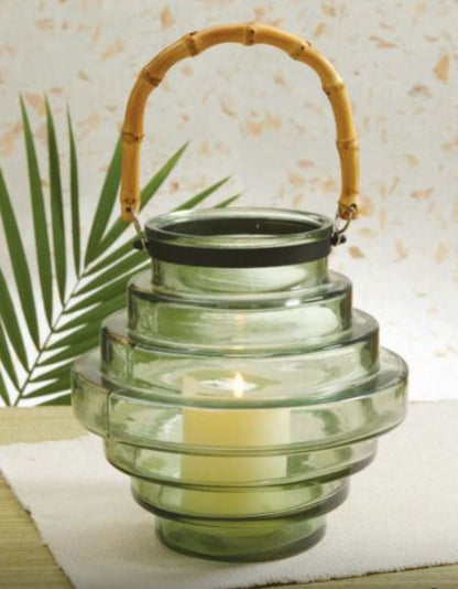 tiered glass lantern with lit candle inside set on a table with greenery in the background.