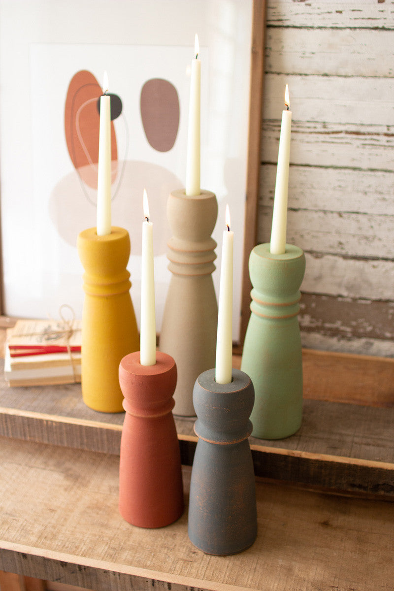 all five colors of the clay tapered candle holders displayed on wooden shelves against a rustic whitewashed slat wall