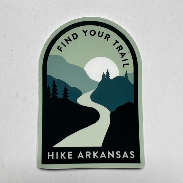 sticker on white background. sticker has graphic of mountain scene in blues and black with sun behind the ridges in background and trail leading toward the sun. "find your trail" is written across the top and "hike arkansas" is along the bottom.