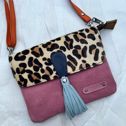 small square bag with dusty rose colored suede body, leopard print flap with light blue tassel and orange strap.