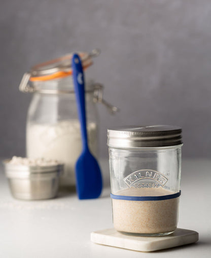 jar of sourdough start on countertop with jar of flour and spatula in background.